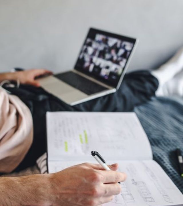 Student taking notes with a notebook during an online class on a laptop, a modern distance learning setup.