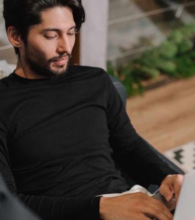 A bearded man in black long sleeves relaxes indoors using a laptop, embodying a modern lifestyle.